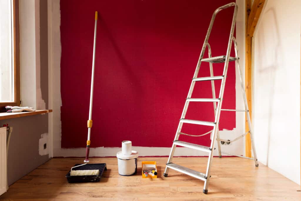 Intérieur en rénovation : mur rouge vif en cours de peinture, échelle métallique, rouleau et pots de peinture blanche sur plancher bois.
