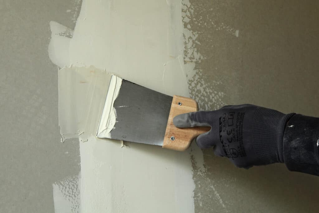 Application d'enduit sur mur avec spatule Main gantée appliquant de l'enduit blanc sur une surface de placoplâtre gris-vert avec une spatule à manche en bois.
