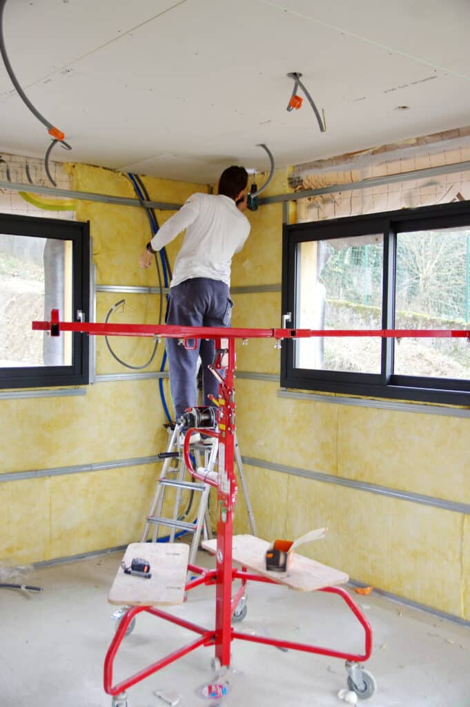Pose de Placo : Chantier Isolation et Cloisonnement Intérieur Ouvrier en pull blanc et pantalon bleu installant des plaques de plâtre sur une ossature métallique, au-dessus de l'isolant jaune, avec un monte-plaques rouge.