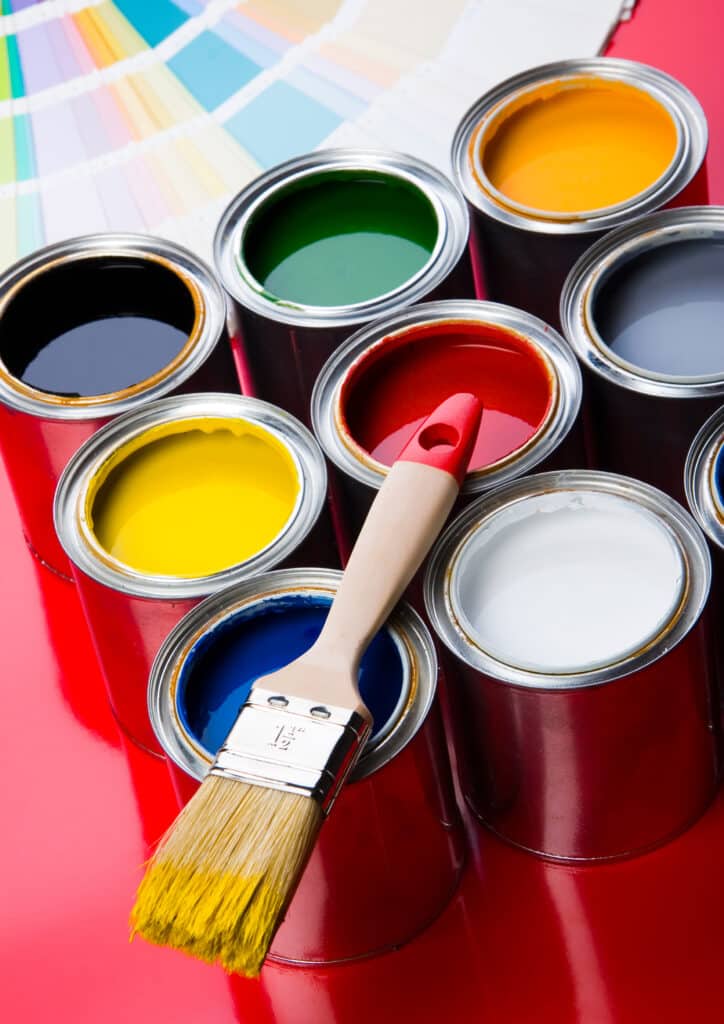 Pots de peinture couleur avec pinceau et nuancier Pinceau de peinture jaune plongé dans de la peinture bleue, entouré de pots de peinture rouge, jaune, verte, blanche, noire et grise, avec nuancier en arrière-plan.